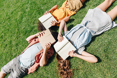 top view of three multicultural schoolkids lying on lawn and covering faces with books