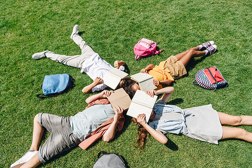 top view of four multicultural schoolkids lying on lawn near backpacks and covering faces with books
