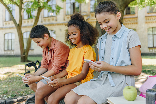 three cute multicultural schoolkids using smartphones while sitting on bench in schoolyard