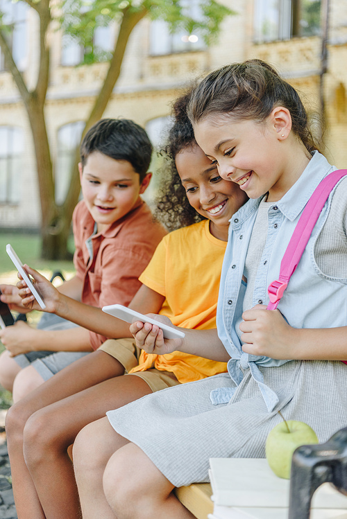 three cheerful multiethnic schoolkids using smartphones while sitting on bench in schoolyard