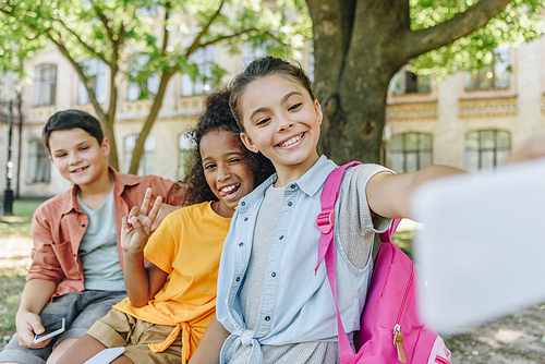 cheerful schoolgirl taking selfie with multicultural friends while sitting on bench in schoolyard