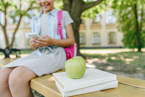cropped view of smiling schoolgirl using smartphone while sitting on bench near books and apple