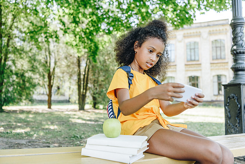 cute african american schoolgirl sitting on bench near books and using smartphone