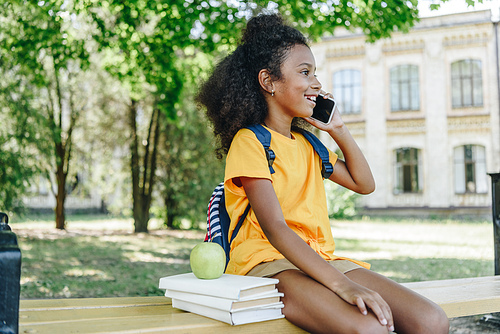 cute african american schoolgirl talking on smartphone while sitting on bench near books and apple
