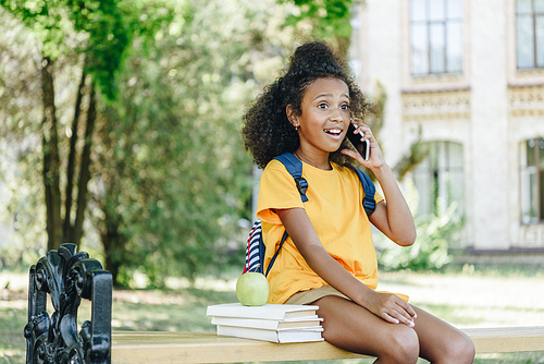 surprised african american girl talking on smartphone while sitting on bench near books and apple