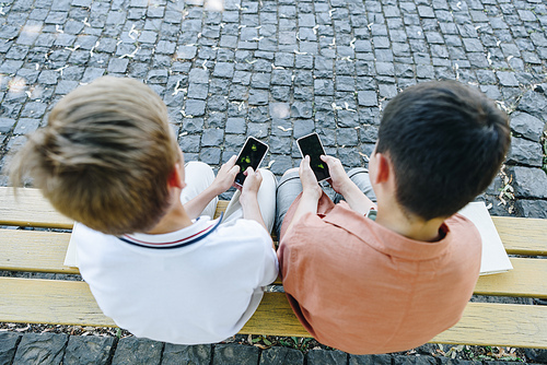 overhead view of two schoolboys sitting on bench and using smartphones