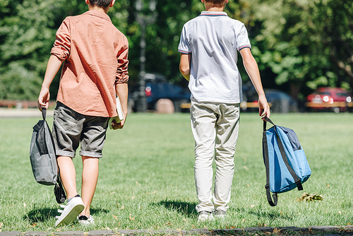 back view of two schoolboys with backpacks walking on lawn in park