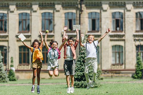 four happy multicultural schoolkids jumping while holding books on lawn in park