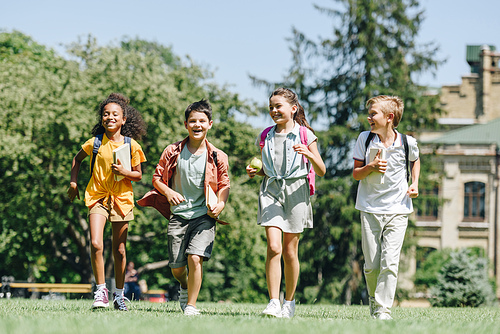 four happy multicultural schoolchildren running on lawn in park together