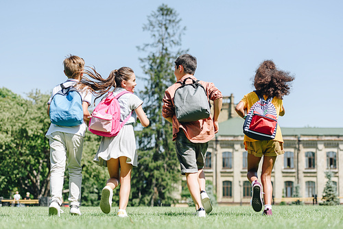 back view of four multiethnic schoolkids with backpacks running on lawn in park