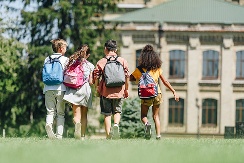 back view of four multicultural schoolkids with backpacks walking on lawn in park