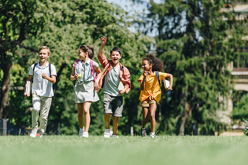 four happy multicultural schoolchildren running on green lawn in park