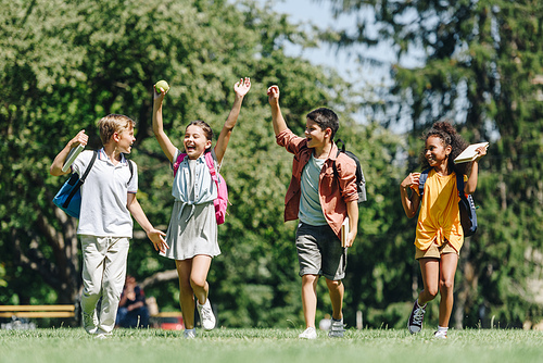 four excited schoolkids gesturing with raised hands while running on lawn in park