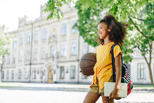 cheerful african american schoolgirl looking away while holding book and ball