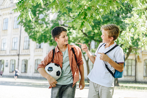 two cheerful schoolboys with backpacks talking while walking in green park