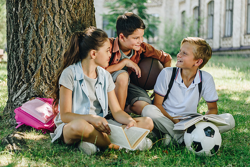 three cheerful schoolkids talking while sitting on lawn under tree