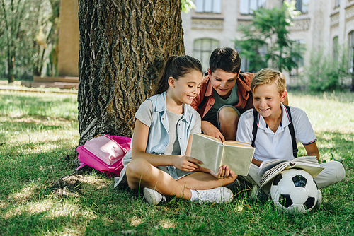three cute classmates reading book and smiling while sitting on lawn under tree