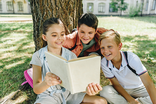 three cheerful schoolkids reading book while sitting under tree in park