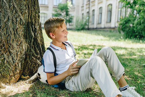 adorable smiling schoolboy sitting on lawn under tree and holding book