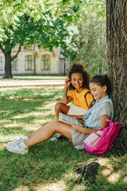 two multicultural schoolgirls reading books while sitting on lawn under tree