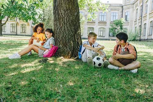 four cute multicultural schoolkids sitting on lawn and tree, speaking and reading books