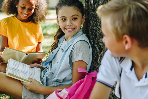 selective focus of three cheerful multicultural classmates speaking while sitting under tree