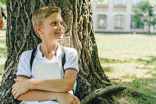adorable schoolboy sitting on lawn under tree, smiling and looking away