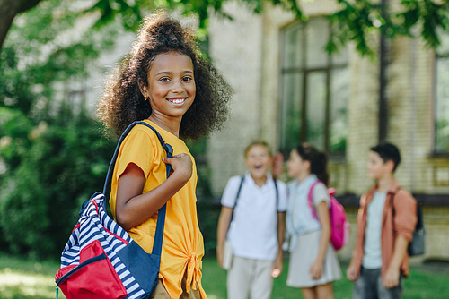 selective focus of cheerful african american schoolgirl with backpack smiling at camera near multiethnic friends
