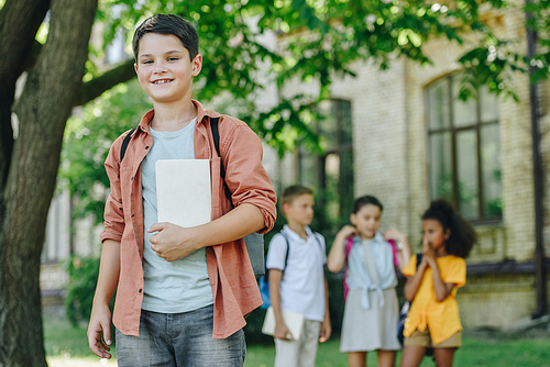 selective focus of adorable schoolboy with book smiling at camera while standing near multicultural friends