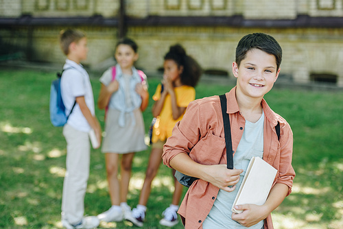 selective focus of smiling schoolboy with book  while standing near multicultural friends