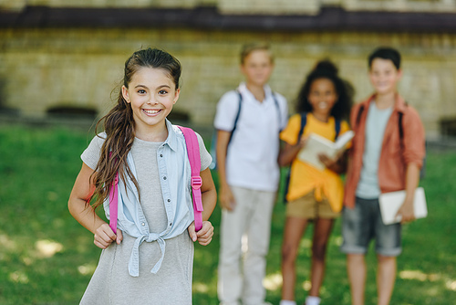 selective focus of adorable schoolgirl smiling at camera while standing near multiethnic friends