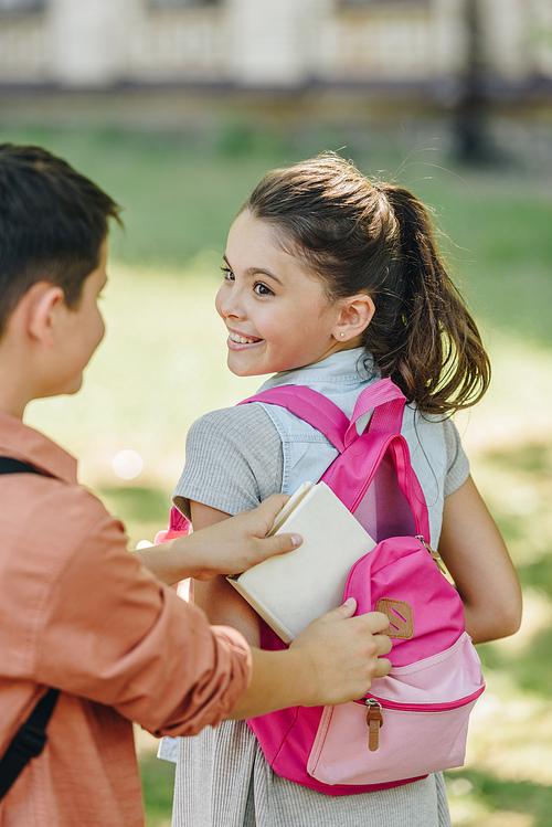 partial view of schoolboy packing book in backpack of cheerful schoolgirl