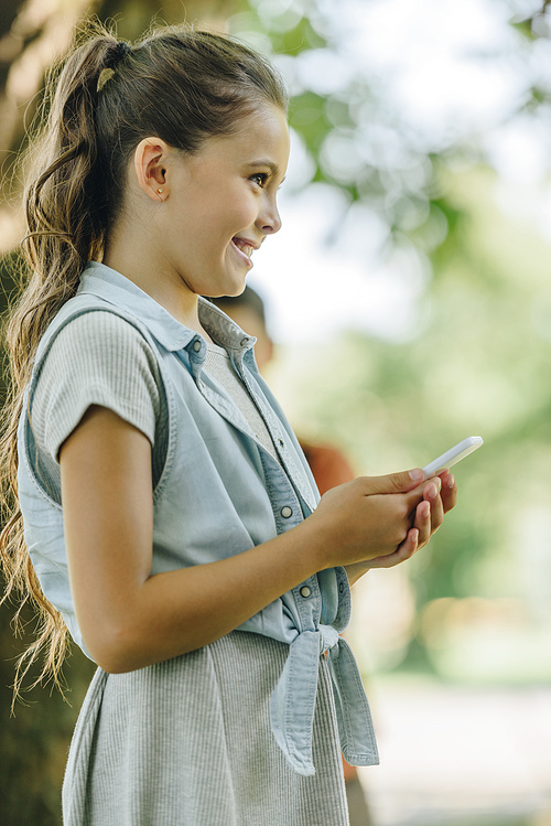 cute, smiling schoolgirl smiling while looking away while using smartphone in park