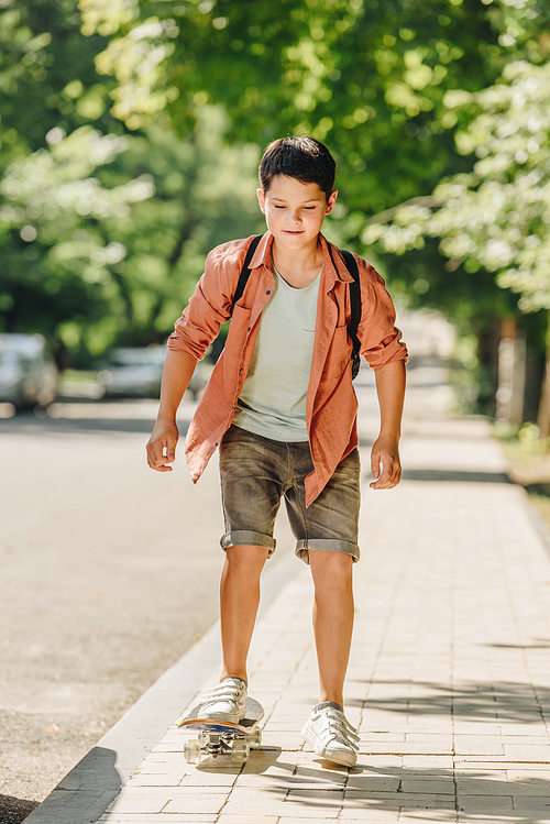 cute schoolboy with backpack riding skateboard on sunny street