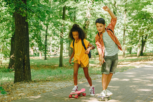 two multicultural schoolkids riding skateboards in green sunny park