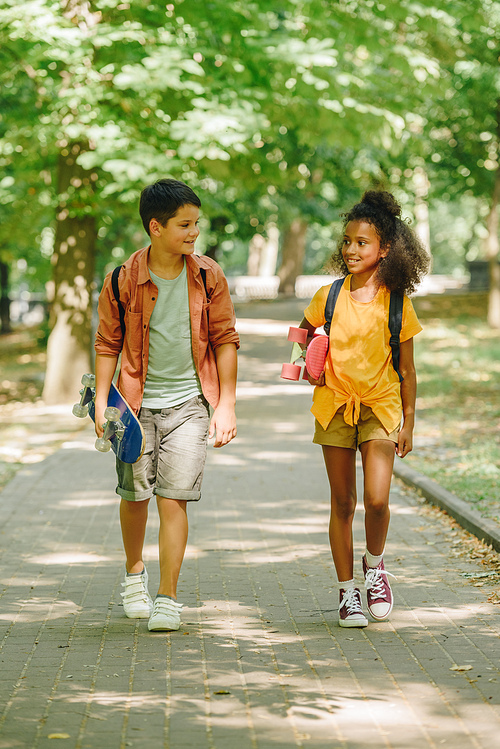 two multicultural schoolkids walking in park while holding skateboards