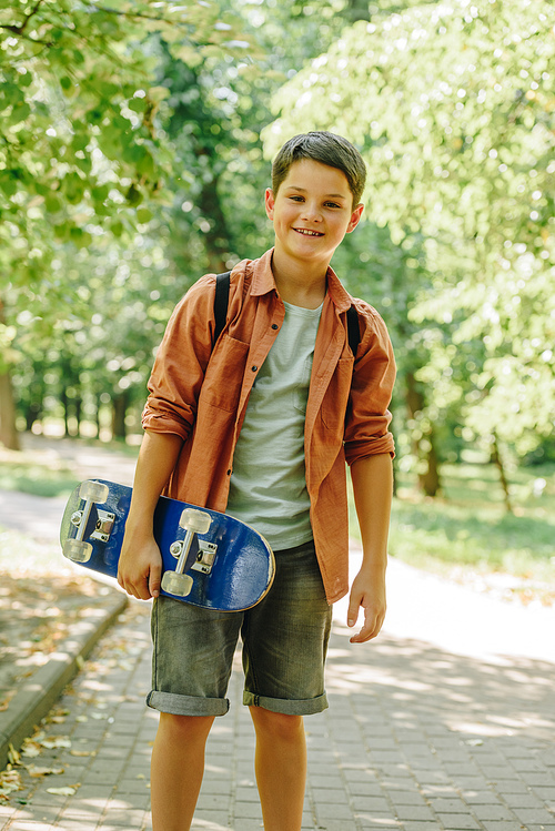 adorable, smiling schoolboy holding skateboard and  in park