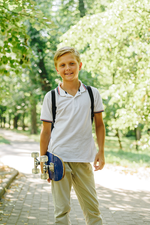 cute, cheerful schoolboy holding skateboard and smiling at camera in park