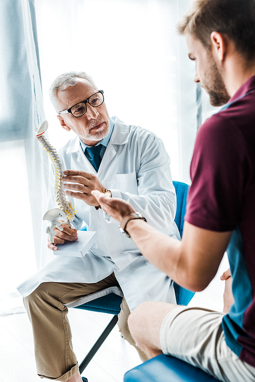 selective focus of bearded doctor in glasses holding spine model near patient gesturing in clinic