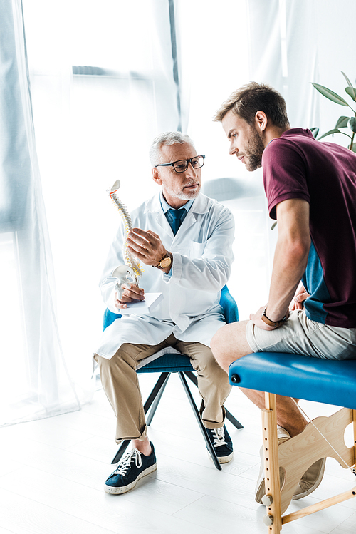 bearded doctor in glasses holding spine model and looking at patient