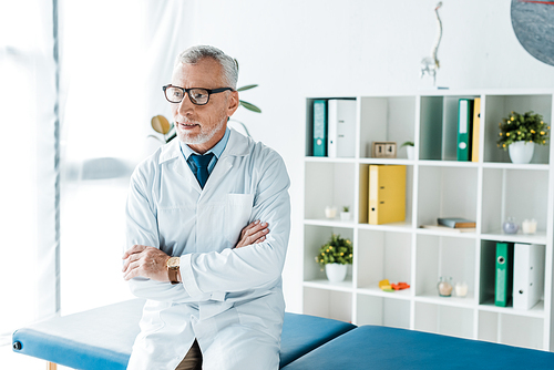 bearded doctor in glasses and white coat sitting on massage table with crossed arms