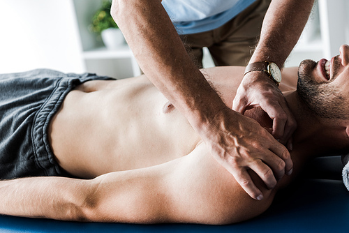 cropped view of chiropractor doing massage to shirtless man with closed eyes suffering pain