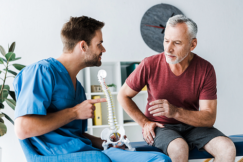 bearded doctor pointing with finger at spine model near patient in clinic