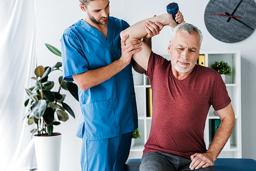 middle aged man exercising with dumbbell near doctor in clinic