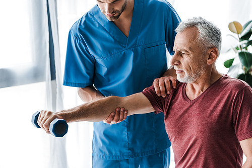 cropped view of doctor standing near mature man holding dumbbell in clinic