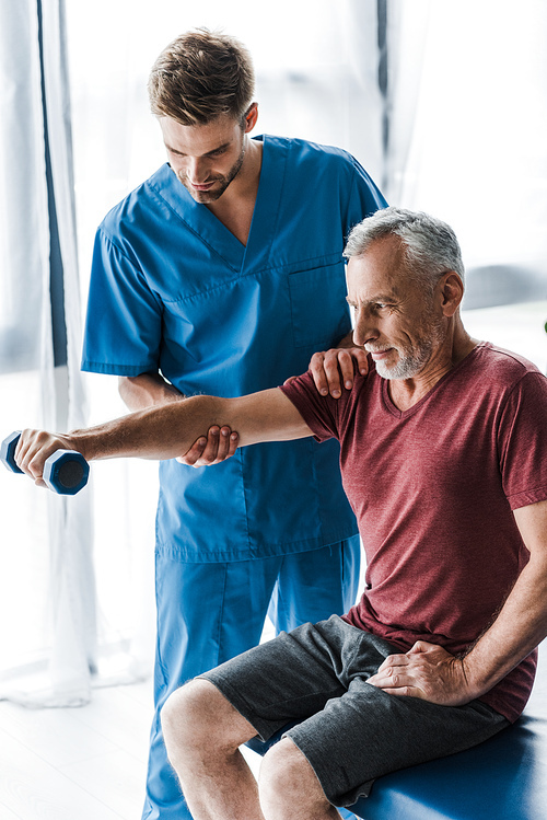 doctor standing near mature man exercising with dumbbell in clinic