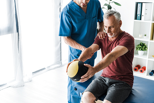 cropped view of doctor standing near mature man exercising with ball