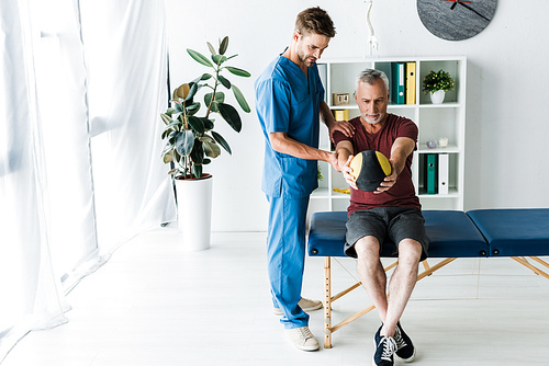 handsome doctor standing near mature man working out with ball