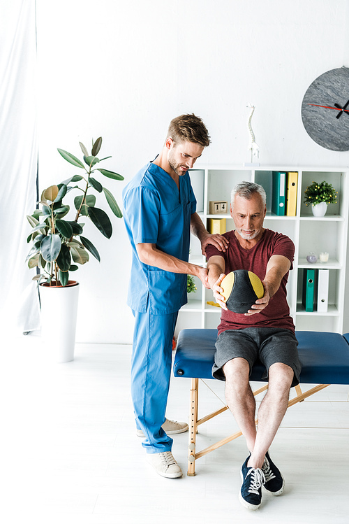 handsome doctor standing near mature man exercising with ball