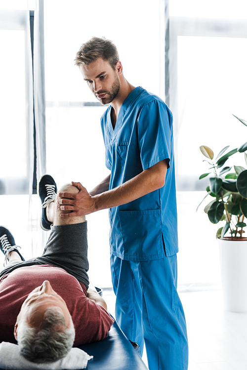 handsome doctor touching leg of mature patient exercising on massage table